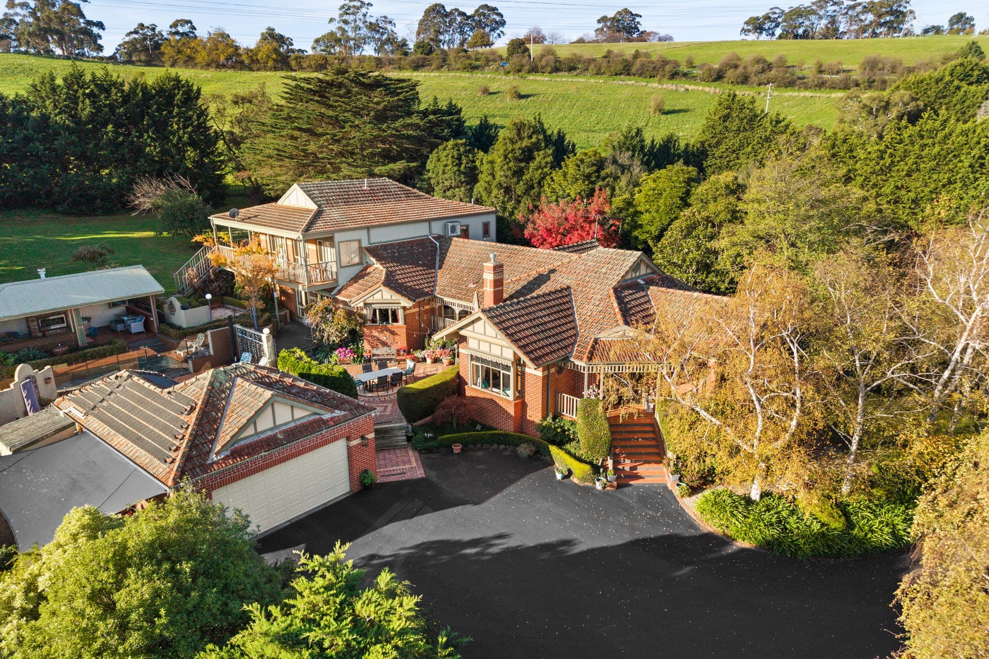 Raised aerial of a large tudor style home in autumn, its roofline clear of the trees and paddocks rising behind it