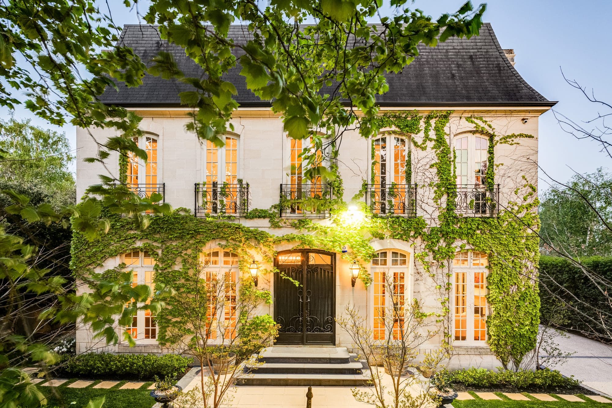 The same facade at dusk, every window lit warm from inside and the entry lantern glowing against the ivy