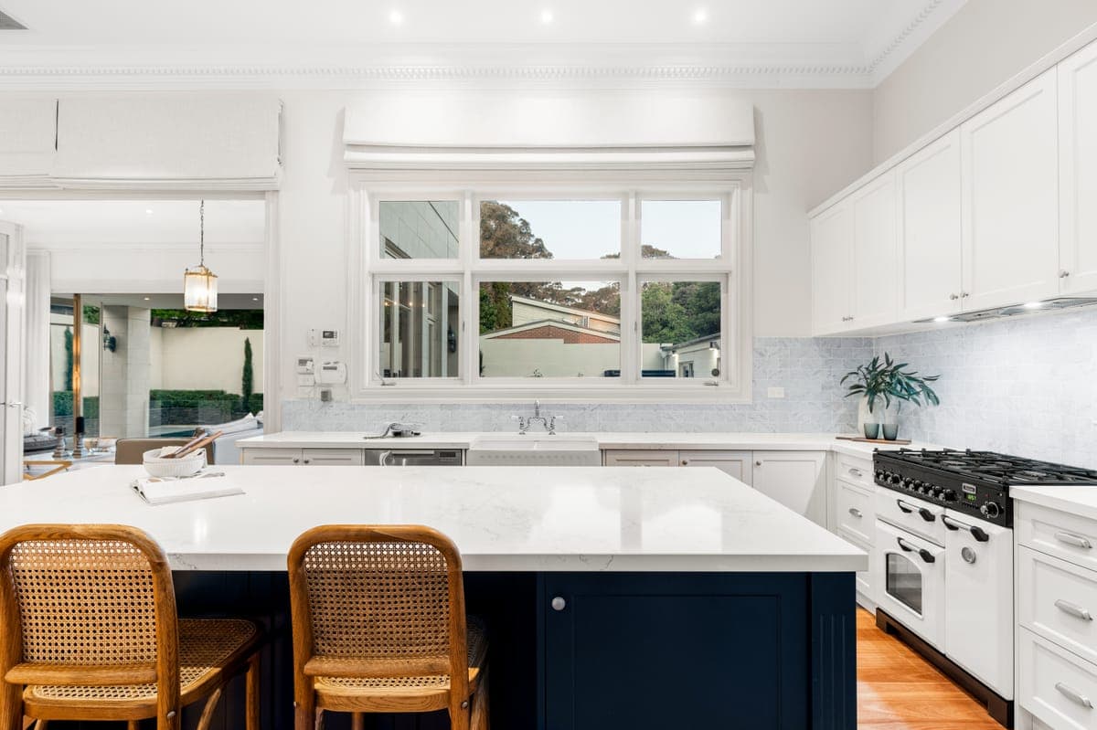 Kitchen with a navy island bench, rattan stools and a window above the sink looking into the garden
